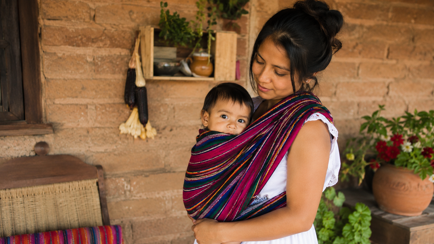 A woman wearing a white dress carries a baby in a colorful woven wrap carrier, standing in front of a rustic brick wall with potted plants.